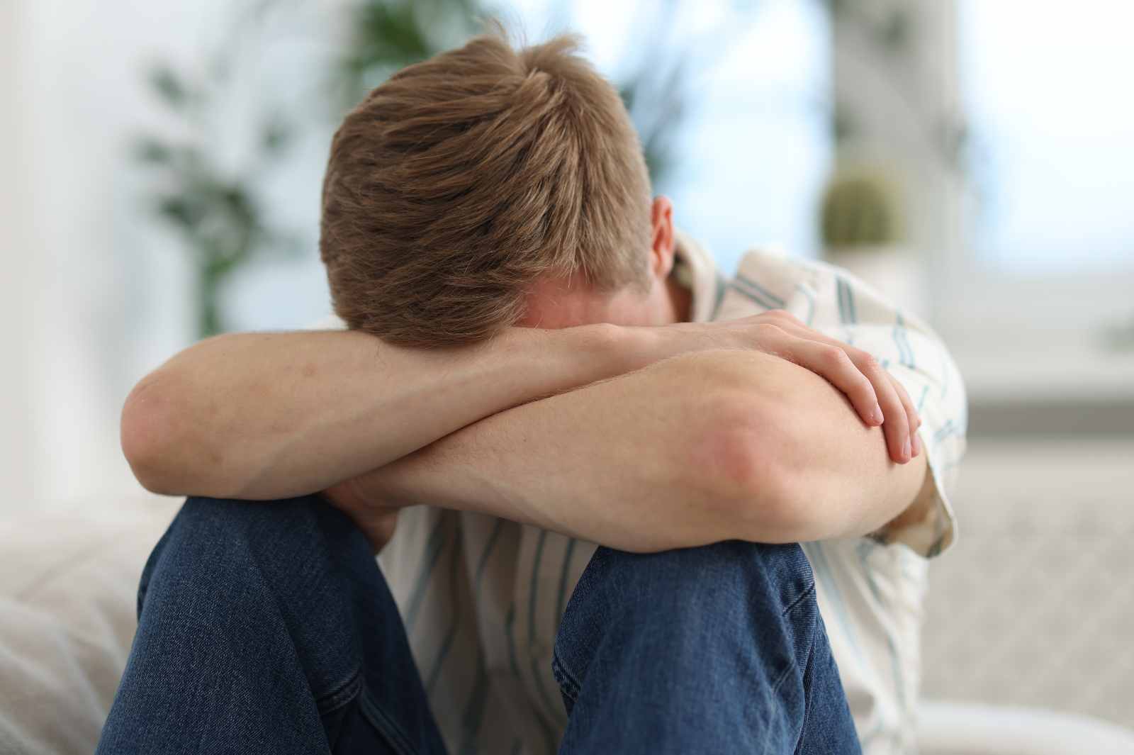 A man sits with his head buried in his crossed arms on his knees.