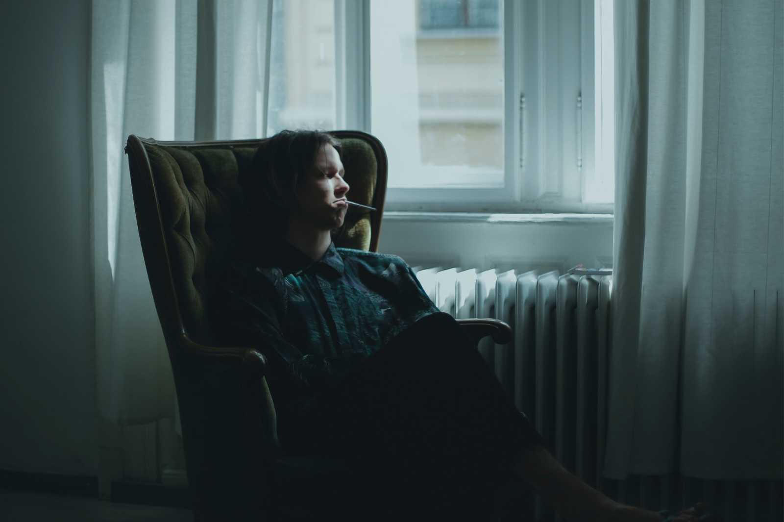 A person sits in a high-back chair by a window with a cigarette.