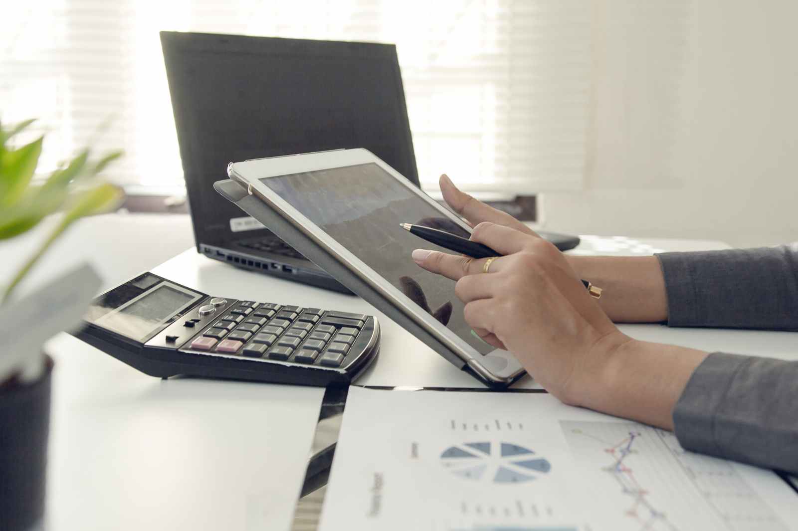 A person uses a stylus on a tablet at a desk with a calculator and charts.