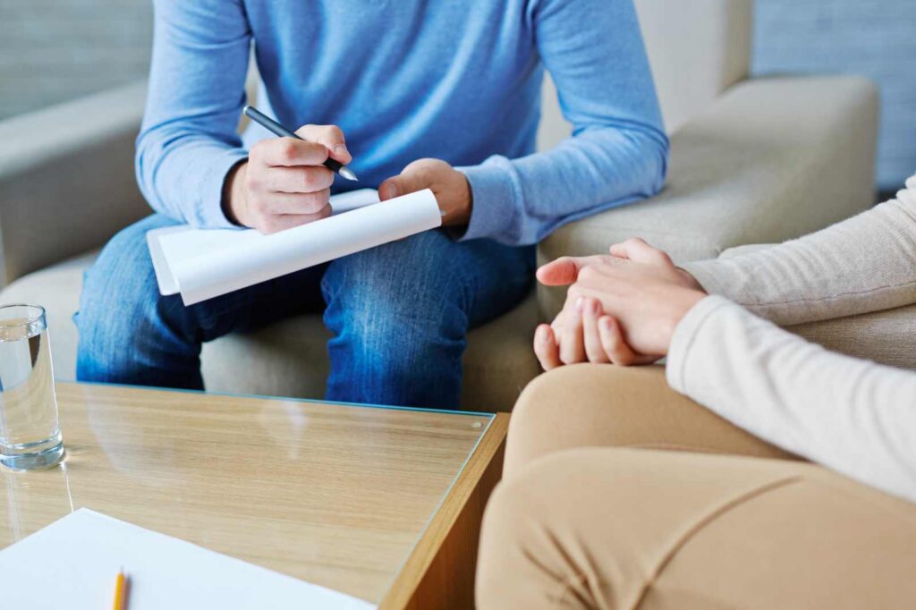 A therapist in a blue shirt takes notes while a client sits with hands clasped.