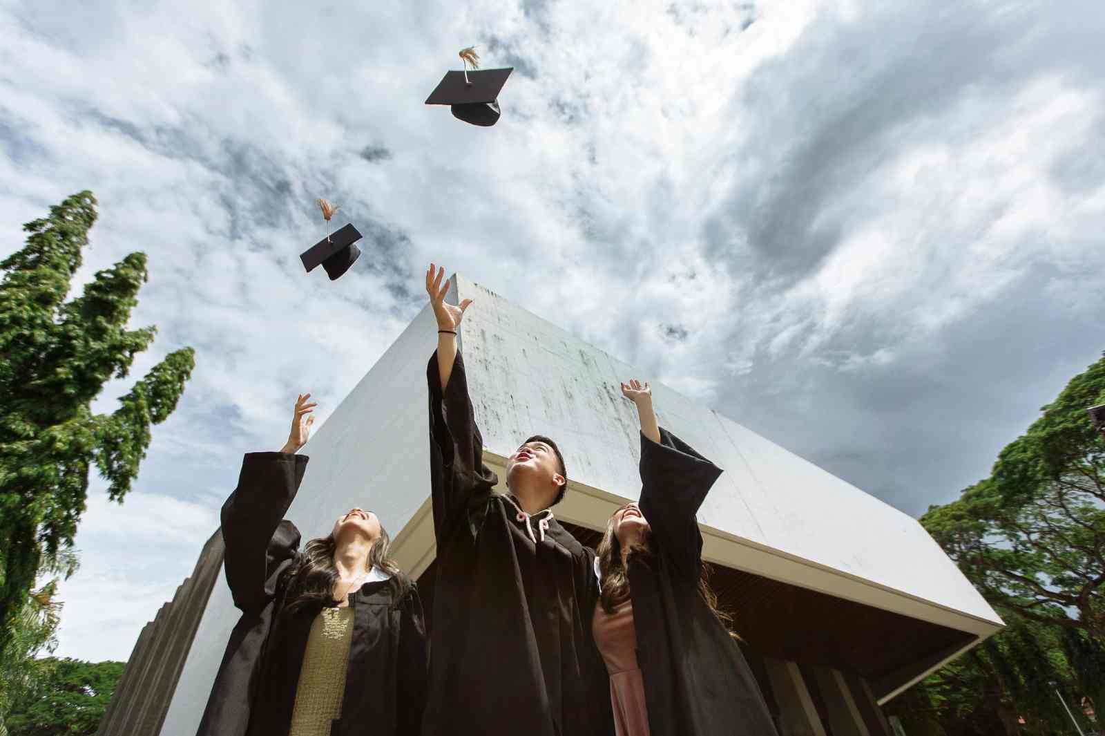 Three graduates in black gowns throw their caps in the air outdoors.