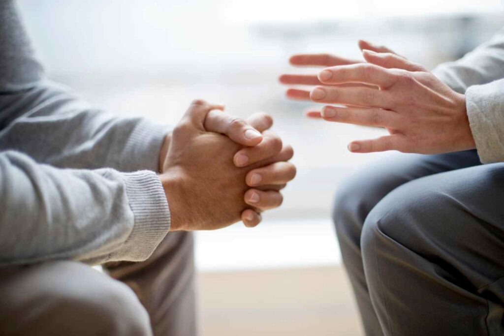 Two people sitting face-to-face, one with hands clasped while the other gestures, suggesting a conversation or counseling session.