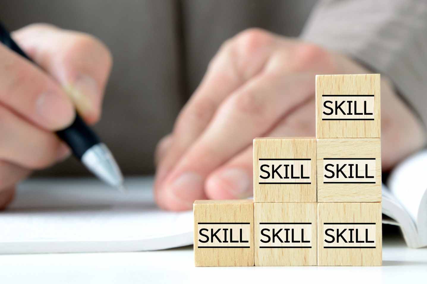 Stacked wooden blocks labeled “skill” with person writing in background.