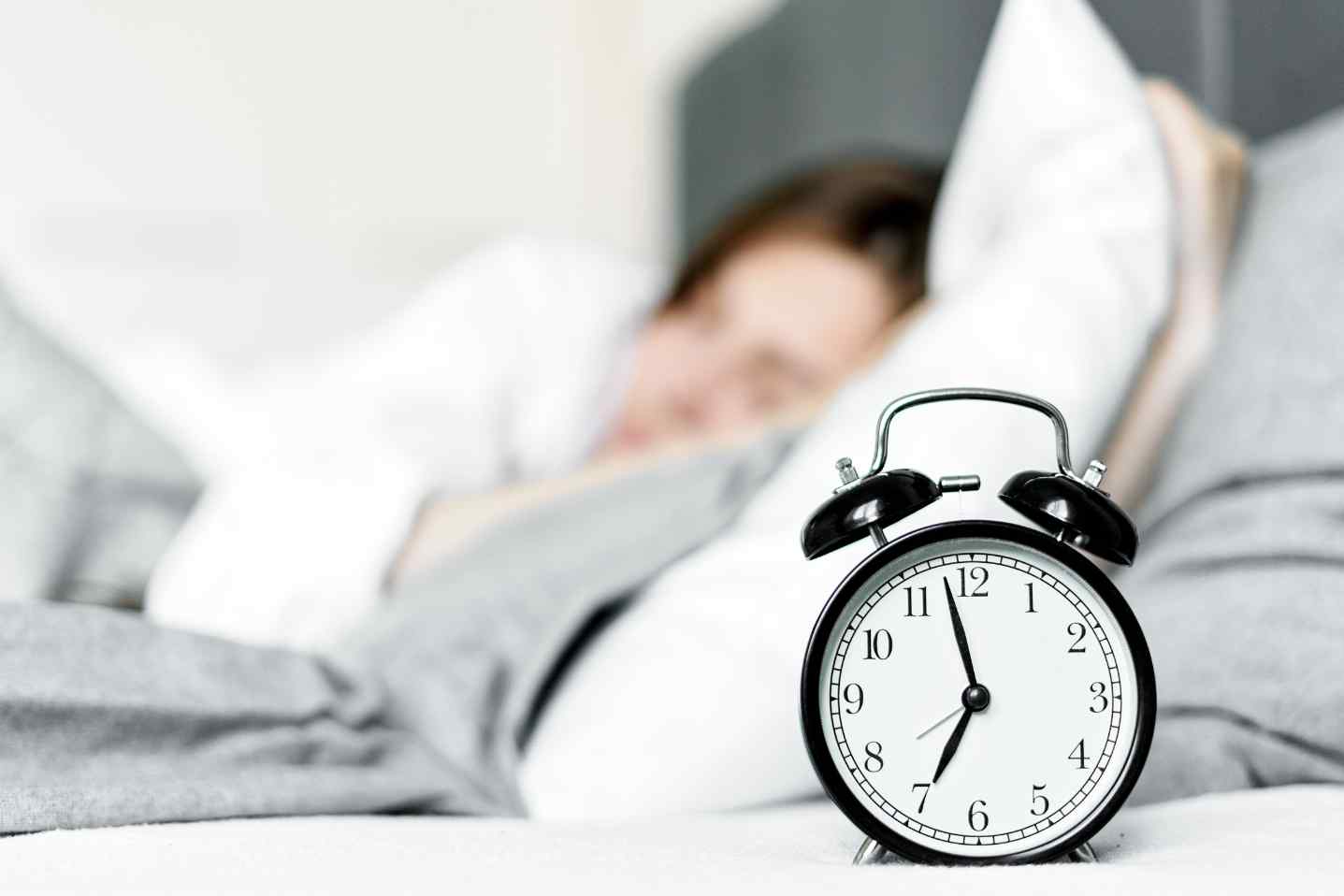 Alarm clock in foreground with person sleeping in bed behind it.