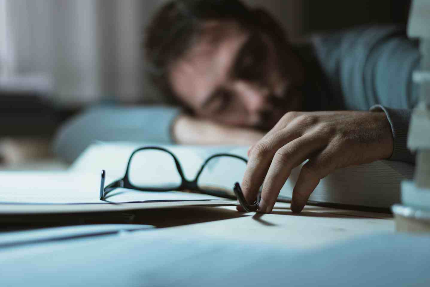 Person lying on desk reaching for glasses, looking tired or exhausted.